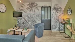 A living room with a sofa, a coffee table, a TV, a wall clock, and a sliding door with a palm tree mural. - Nice apartment in villa for 5 people with WIFI, TV and terrace (Le Magouër)