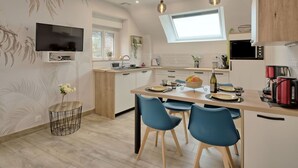 A modern kitchen with a central island, a dining area with blue chairs, a TV mounted on the wall, and a window providing natural light.