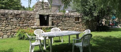 A stone wall with a chimney, a round table, and four chairs in a grassy outdoor area.