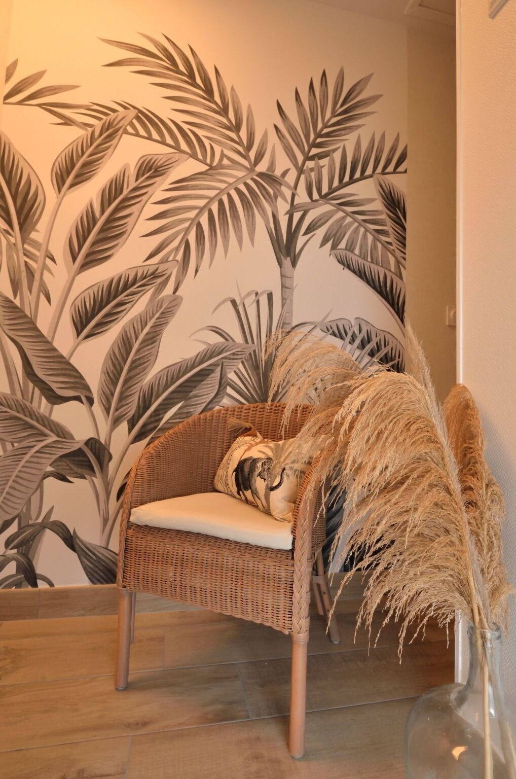A wicker chair with a cushion in front of a wall with a tropical leaf pattern.