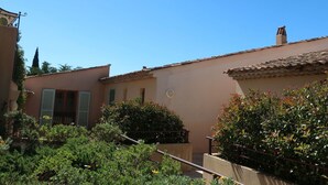 A traditional Mediterranean-style building with terracotta tiled roofs, a small garden area with various shrubs and flowers, and a clear blue sky. - Lovely apartment for 6 guests with WIFI, A/C, TV, terrace and panoramic view (Grimaud)
