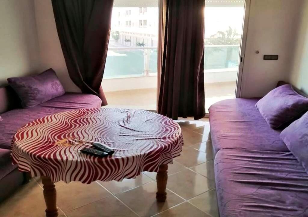 Una habitación con una mesa redonda cubierta con un mantel a rayas de cebra, dos sofás morados y vistas a un edificio y una masa de agua a través de la ventana.