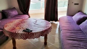 A room with a round table covered in a zebra-striped tablecloth, two purple sofas, and a view of a building and a body of water through the window. - Residence Yasmine Mdiq 4691 (Mdiq)