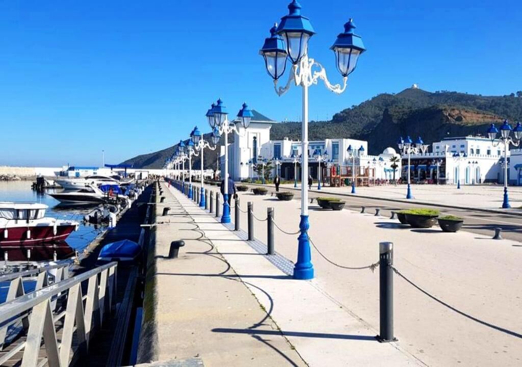 A coastal promenade with a row of white buildings, blue street lamps, and boats docked along the water.