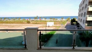 A view from a balcony overlooking a beach with a clear sky, palm trees, and a building in the background. - Residence Yasmine Mdiq 4692 (Mdiq)