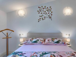 Un dormitorio con una cama, almohadas estampadas, lámparas de noche, una lámpara de pared y una silla de madera.