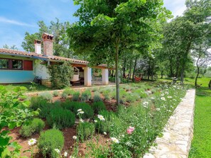 Une maison avec un toit en tuiles, une cheminée en pierre et un jardin où poussent différentes plantes et fleurs.