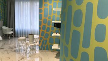 Una habitación de hotel moderna con piso de mármol, una zona de estar con una silla blanca y una mesita redonda pequeña, y un ventanal con cortinas de tela ligera.
