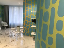 Een moderne hotelkamer met een marmeren vloer, een zithoek met een witte stoel en een klein rond tafeltje, en een groot raam met lichte gordijnen.