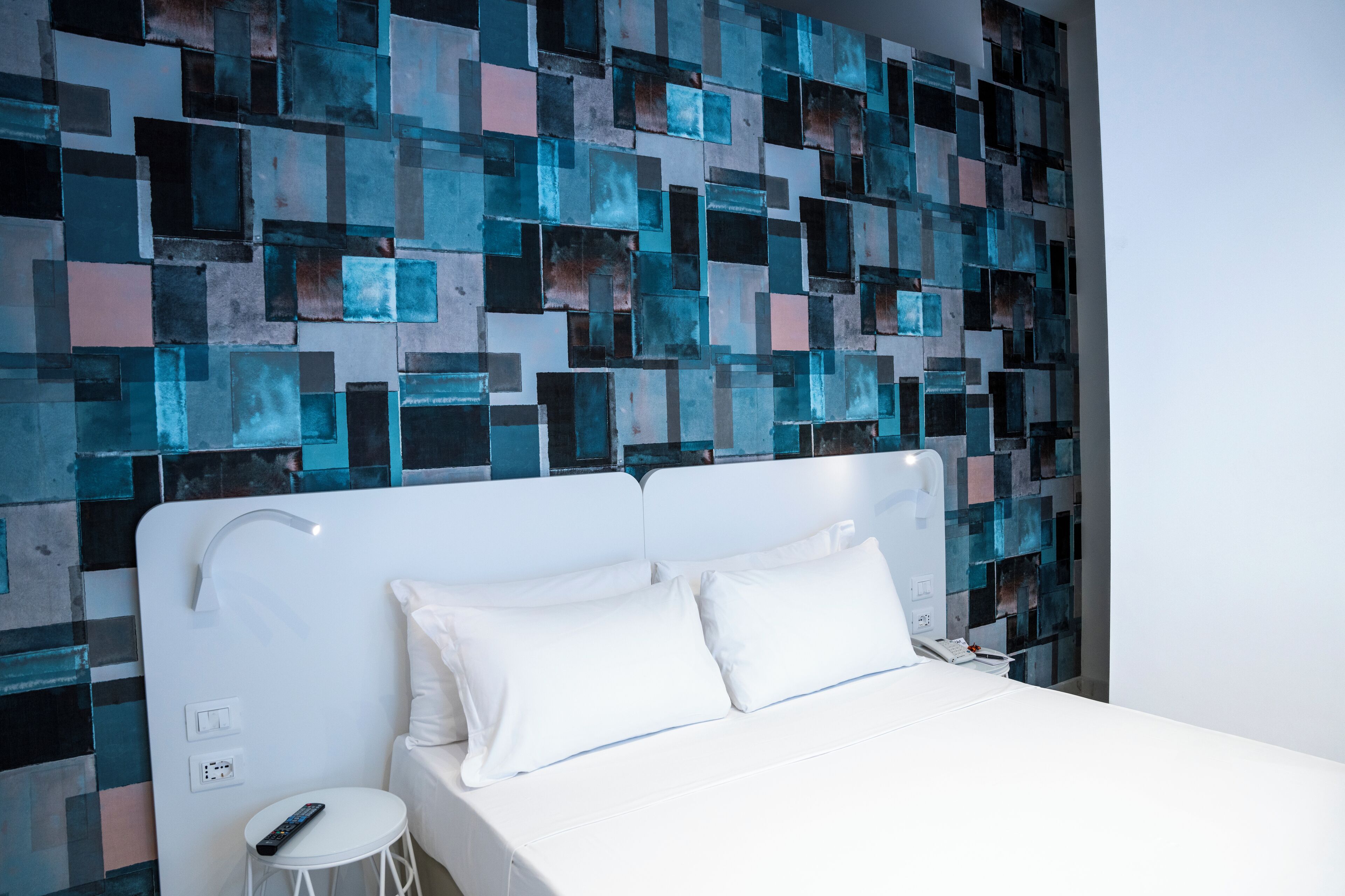 A modern hotel room with a white bed, a small white stool, and a wall with a geometric mosaic design.