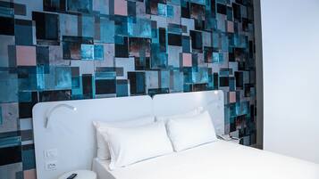 A modern hotel room with a white bed, a small white stool, and a wall with a geometric mosaic design.