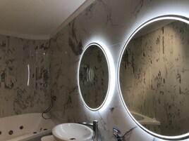 Un bagno con parete in marmo, uno specchio rotondo illuminato con luci soffuse, un lavabo bianco e una vasca da bagno.