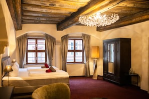 A hotel room with a bed, two windows with curtains, a chandelier, a wardrobe, and a lamp.