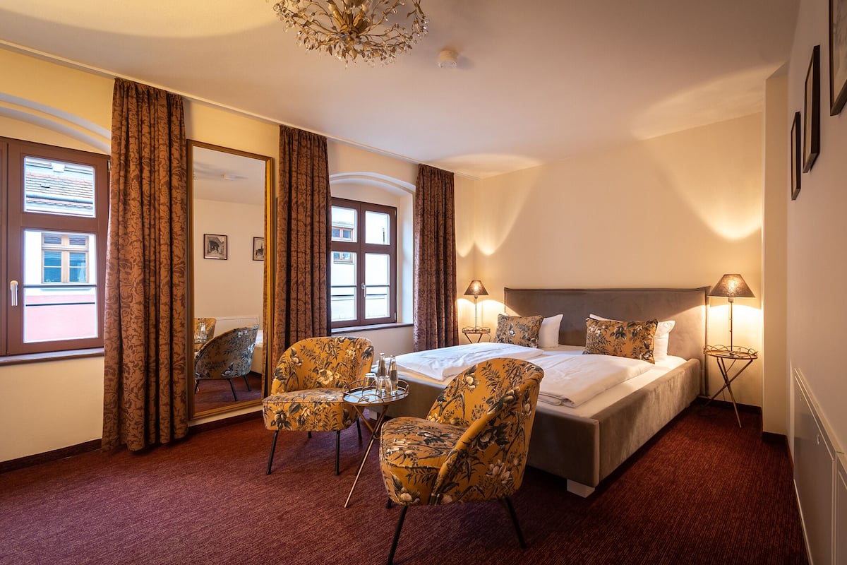 Quarto de hotel com cama, duas cadeiras, mesa pequena, abajur e janela com cortinas.