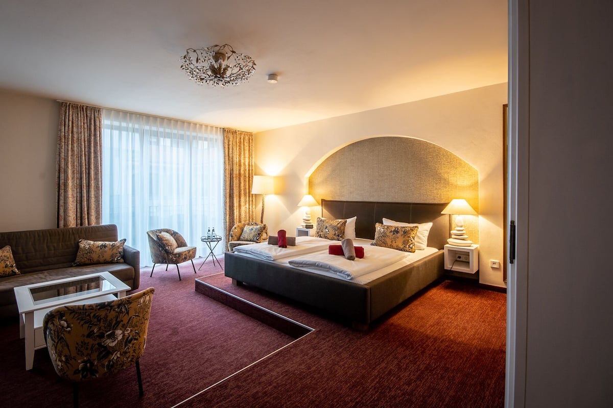Quarto de hotel com uma cama grande, um sofá, uma cadeira, uma mesinha e uma luminária decorativa no teto.