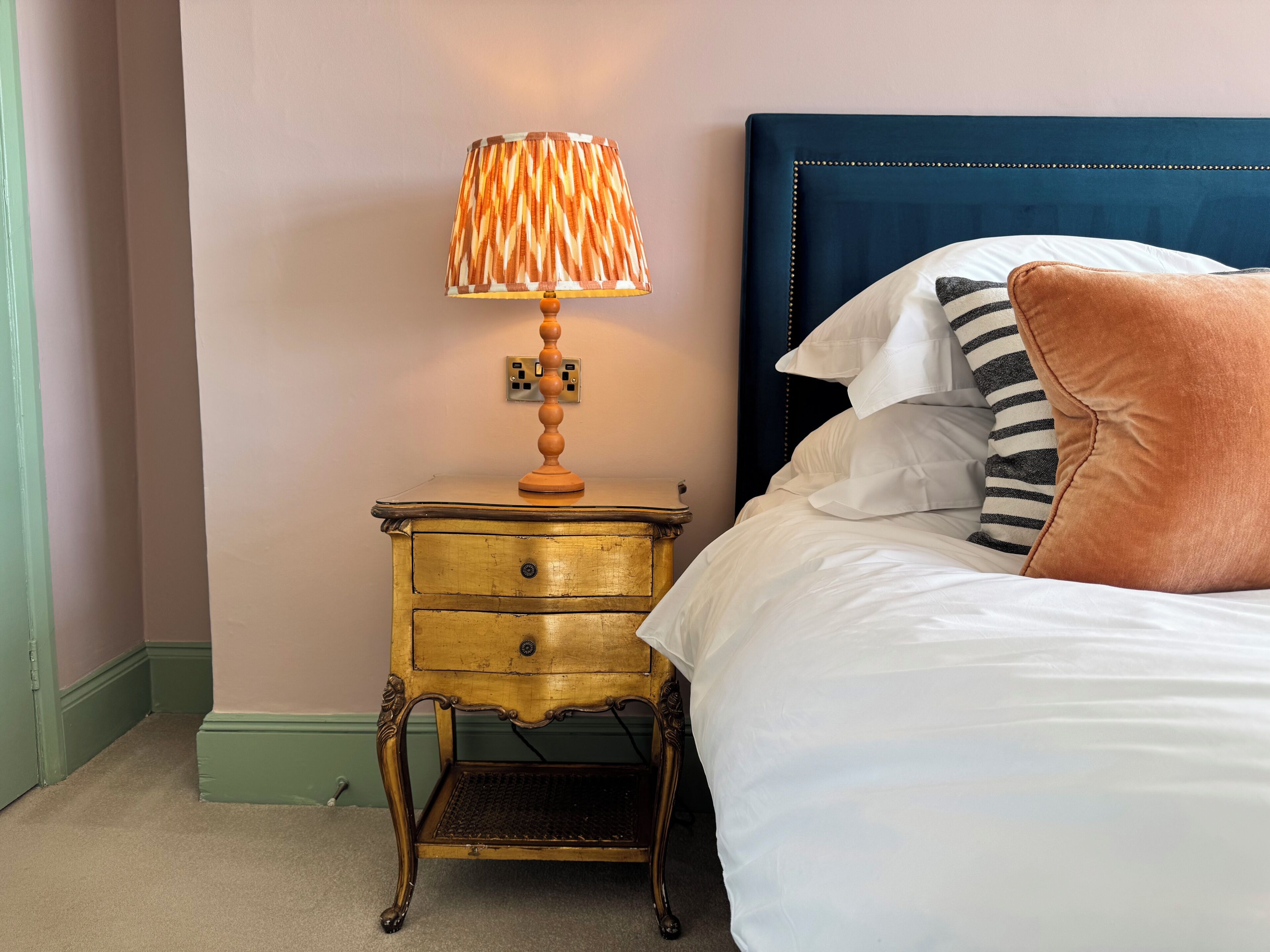 Un lit avec une tête de lit bleue, une lampe de chevet avec un abat-jour à motifs et une petite table de chevet en bois dotée de tiroirs.