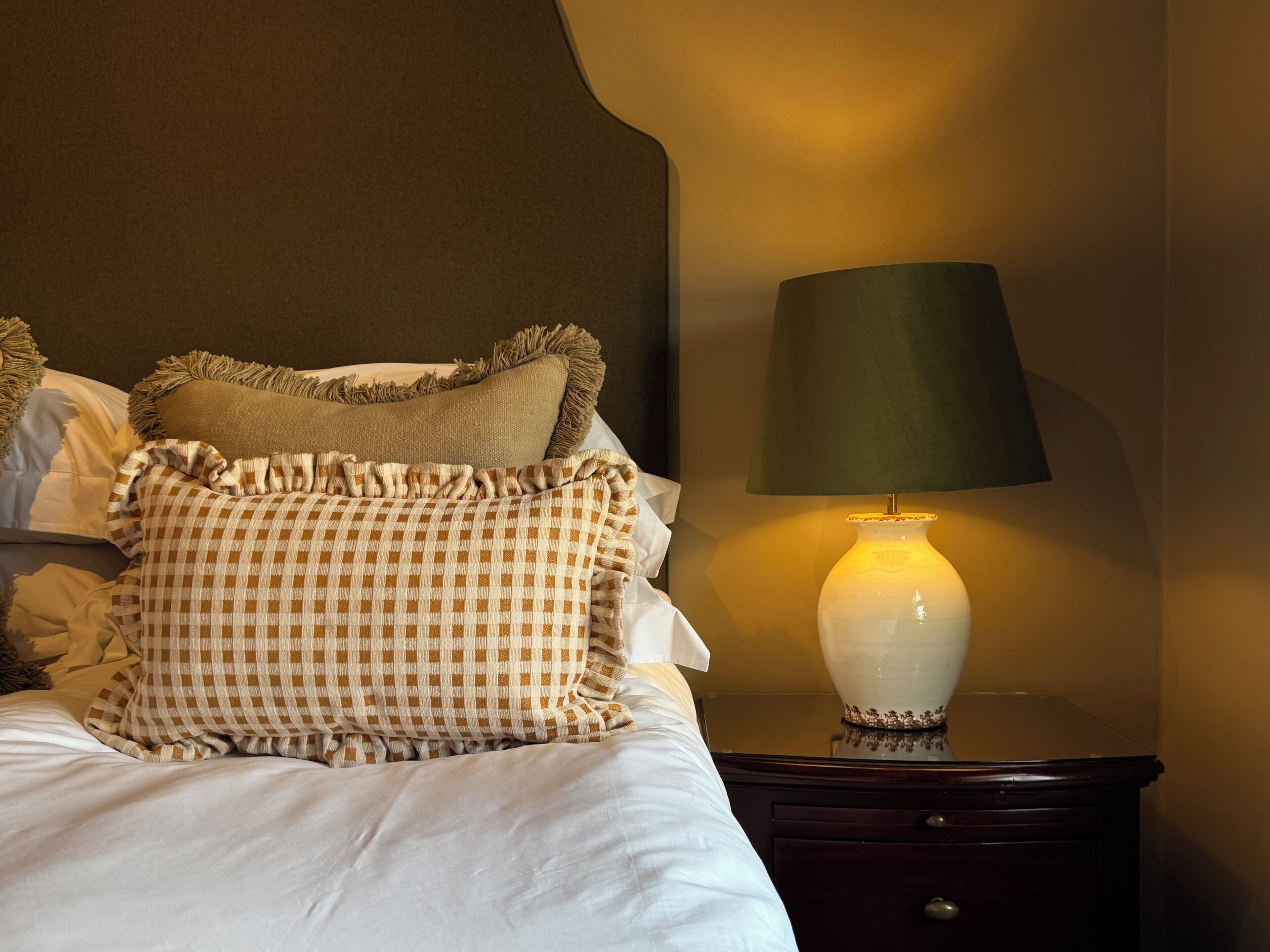 A neatly made bed with a checkered blanket and pillows, a bedside lamp, and a dark nightstand.