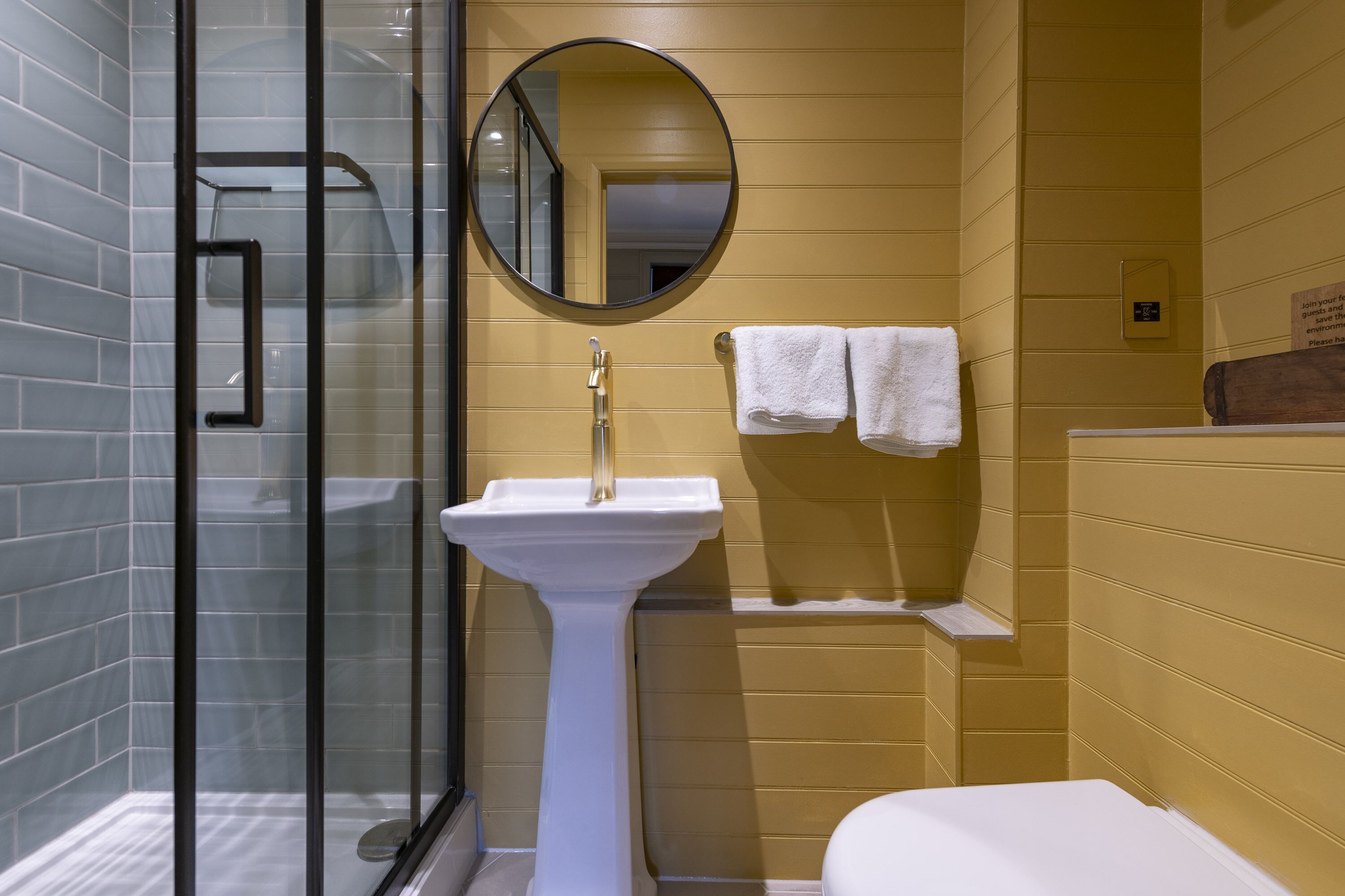 A bathroom with a glass shower enclosure, a pedestal sink, and a round mirror.