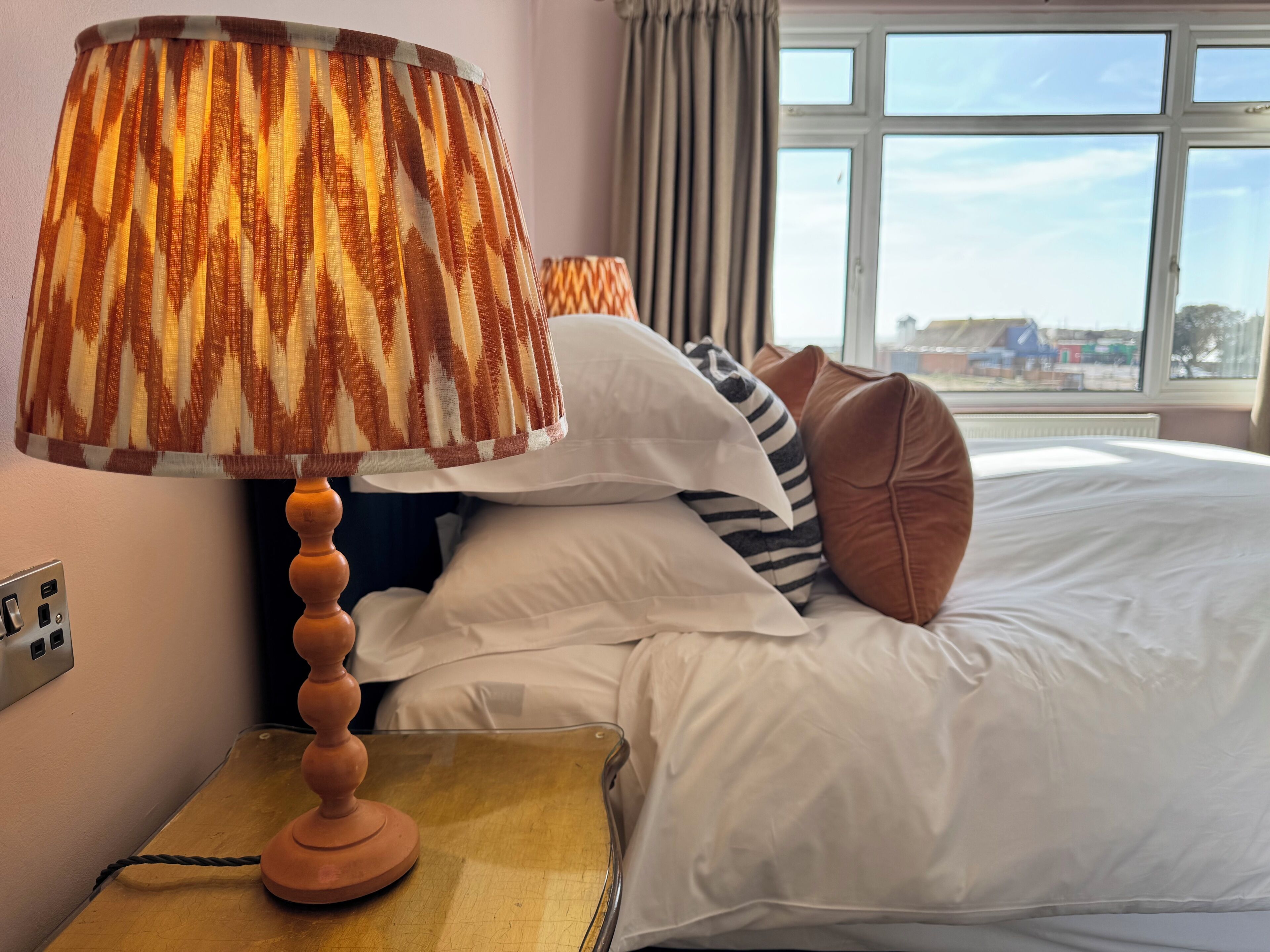 Habitación de hotel con cama, almohadas, lámpara, mesita de noche y ventana con vistas.