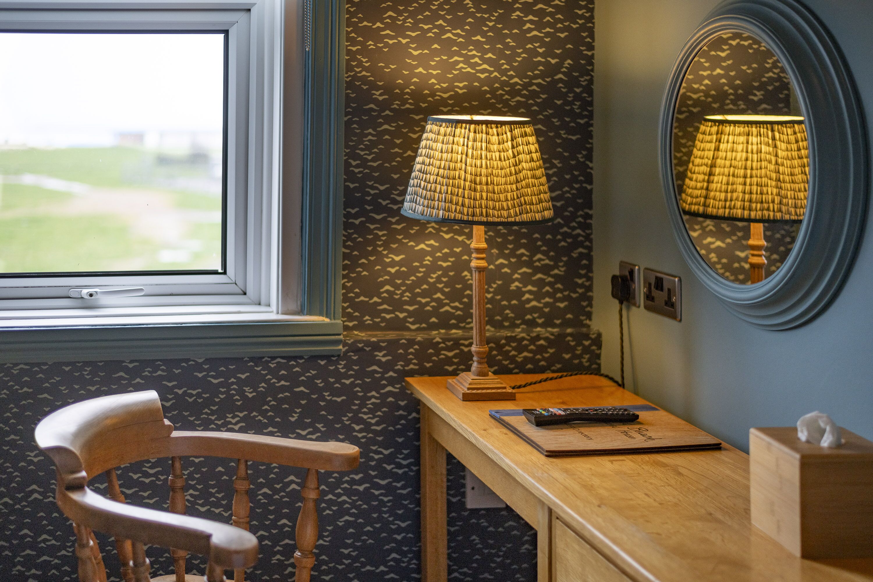 A room with a wooden chair, a desk with a lamp, a remote control, and a tissue box.