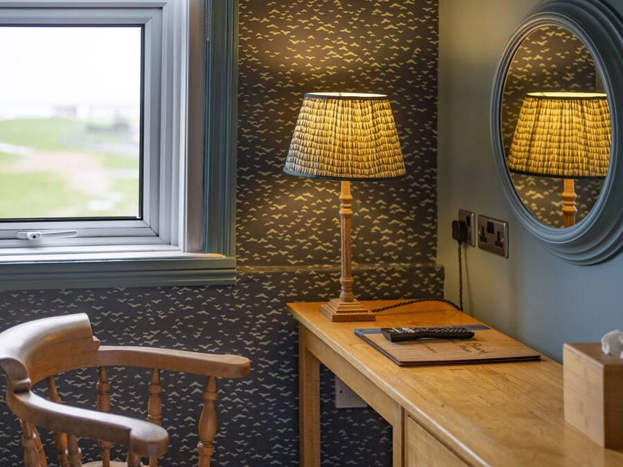 A room with a wooden chair, a desk with a lamp, a remote control, and a tissue box.