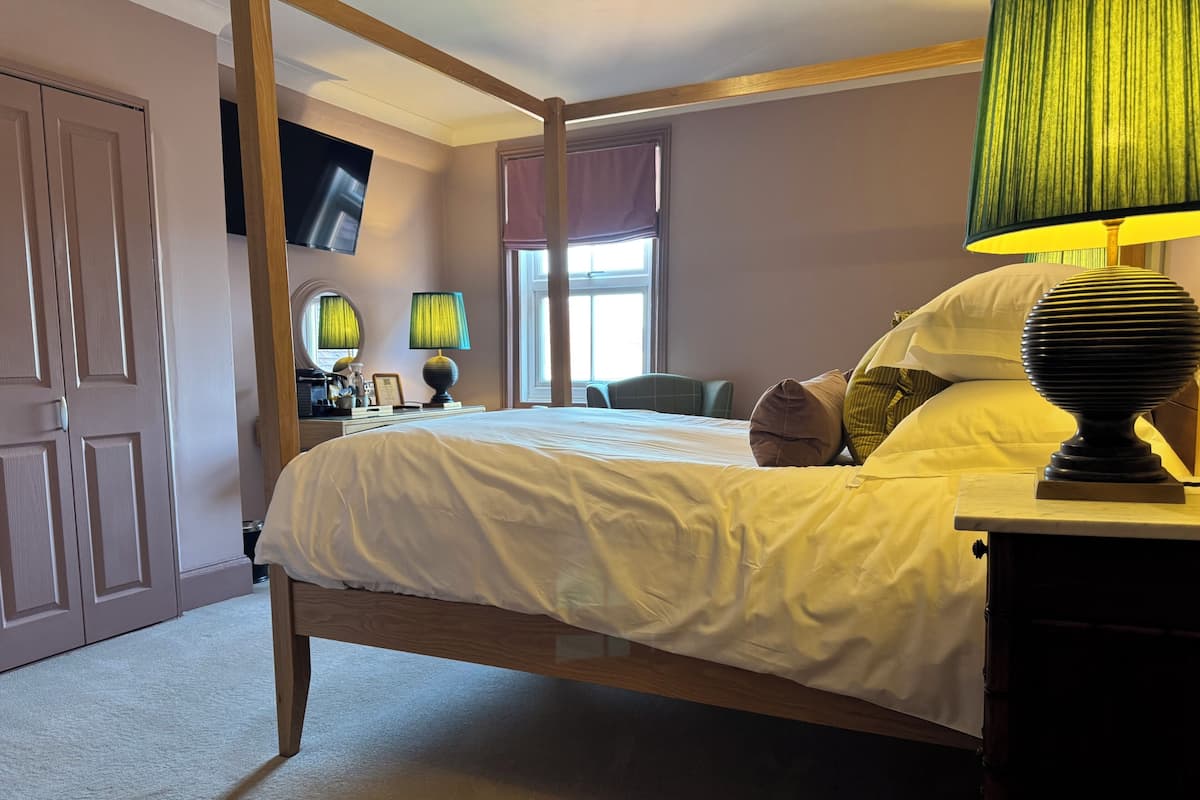A bedroom with a four-poster bed, a bedside table with a lamp, a desk with a computer, a window with blinds, and a wardrobe.