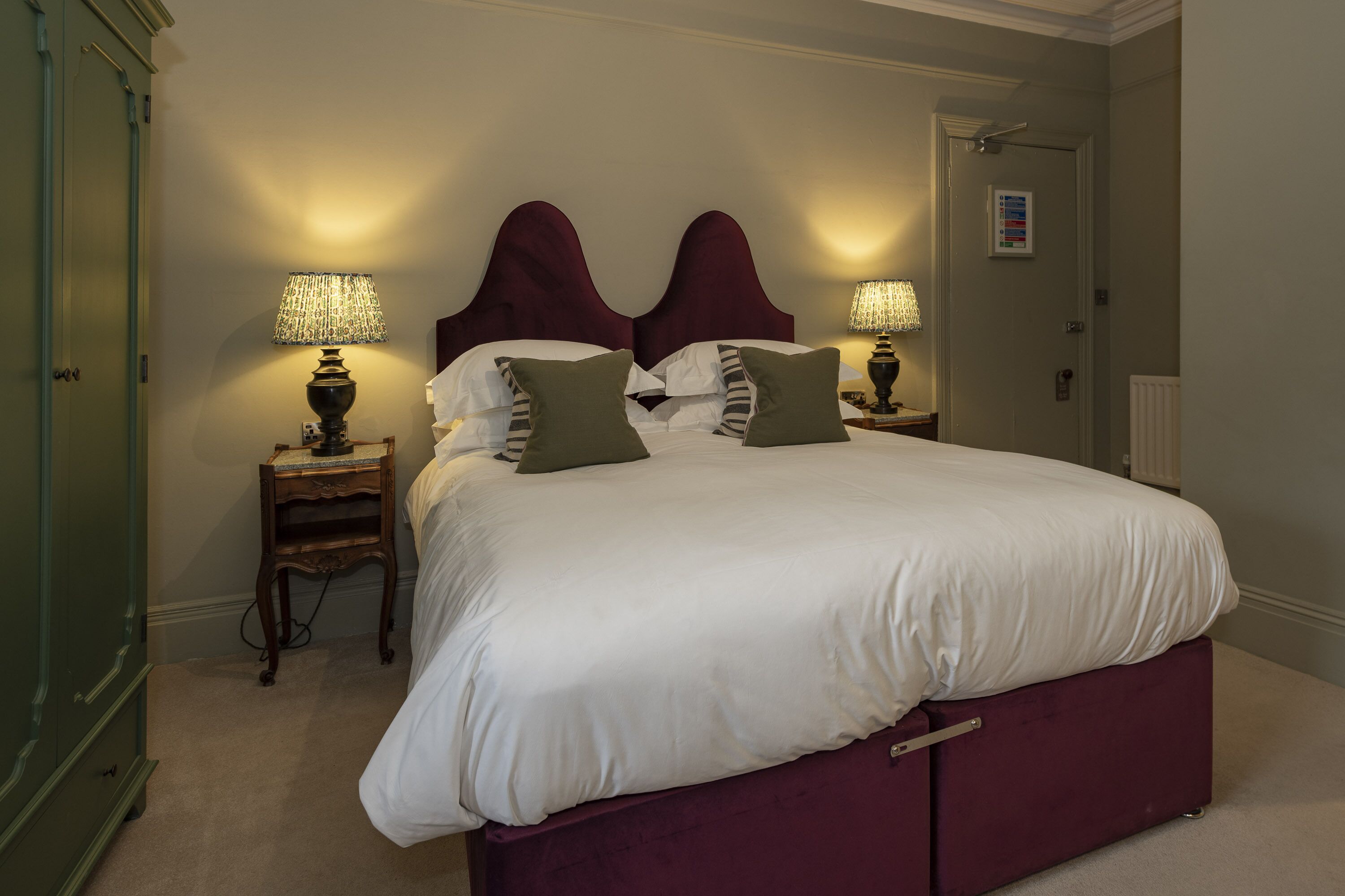 A neatly made bed with a white duvet and green pillows, flanked by two bedside tables with lamps, in a room with a green wardrobe and a door with a sign.
