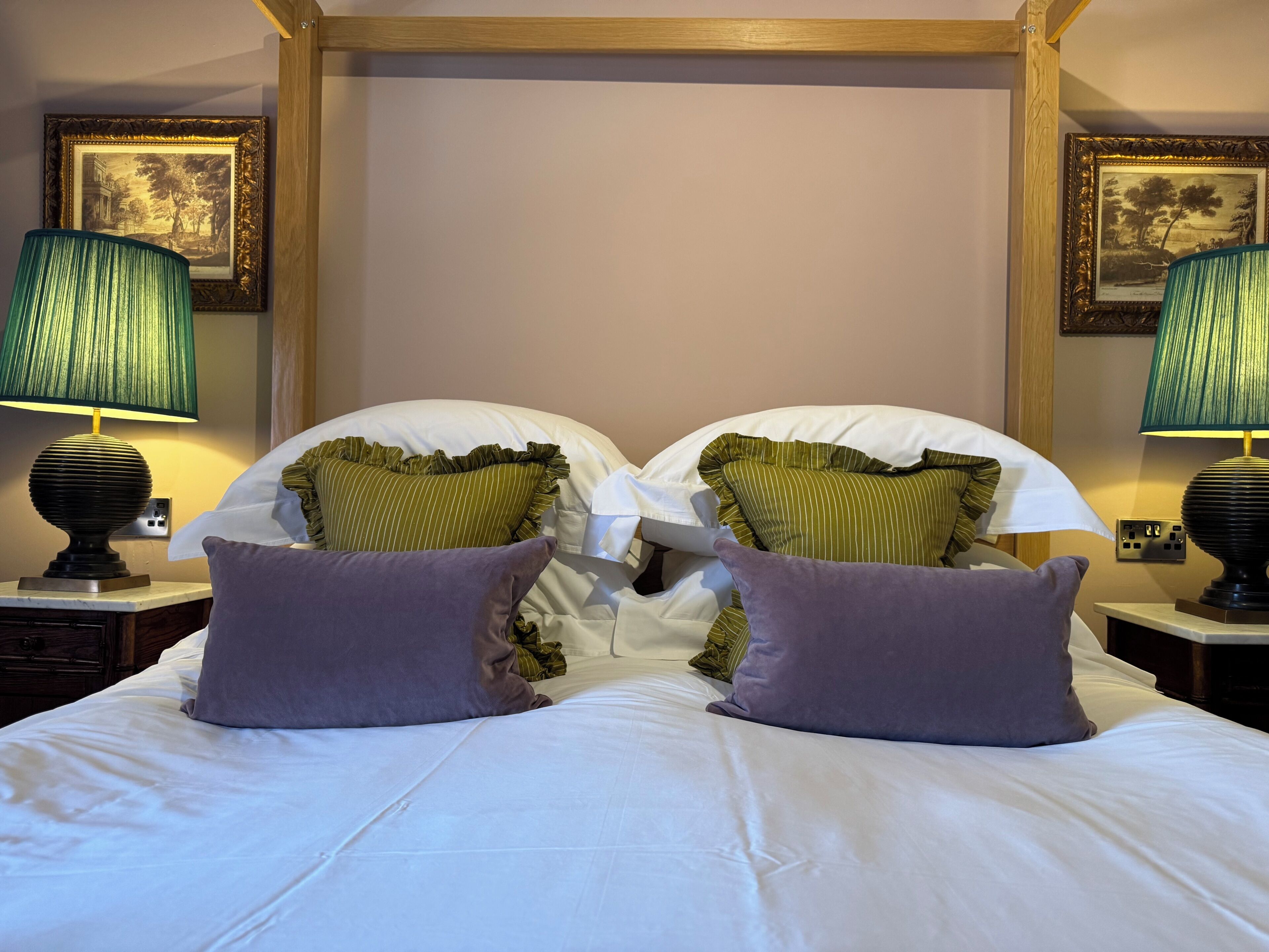 Una cama bien hecha con sábanas blancas, dos almohadas a rayas verdes y una almohada morada. Hay dos lámparas con pantallas verdes a cada lado de la cama.