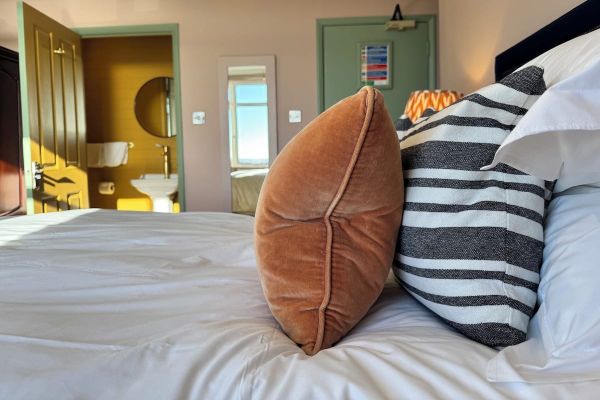 A hotel room with a bed, pillows, a door, a mirror, and a window.