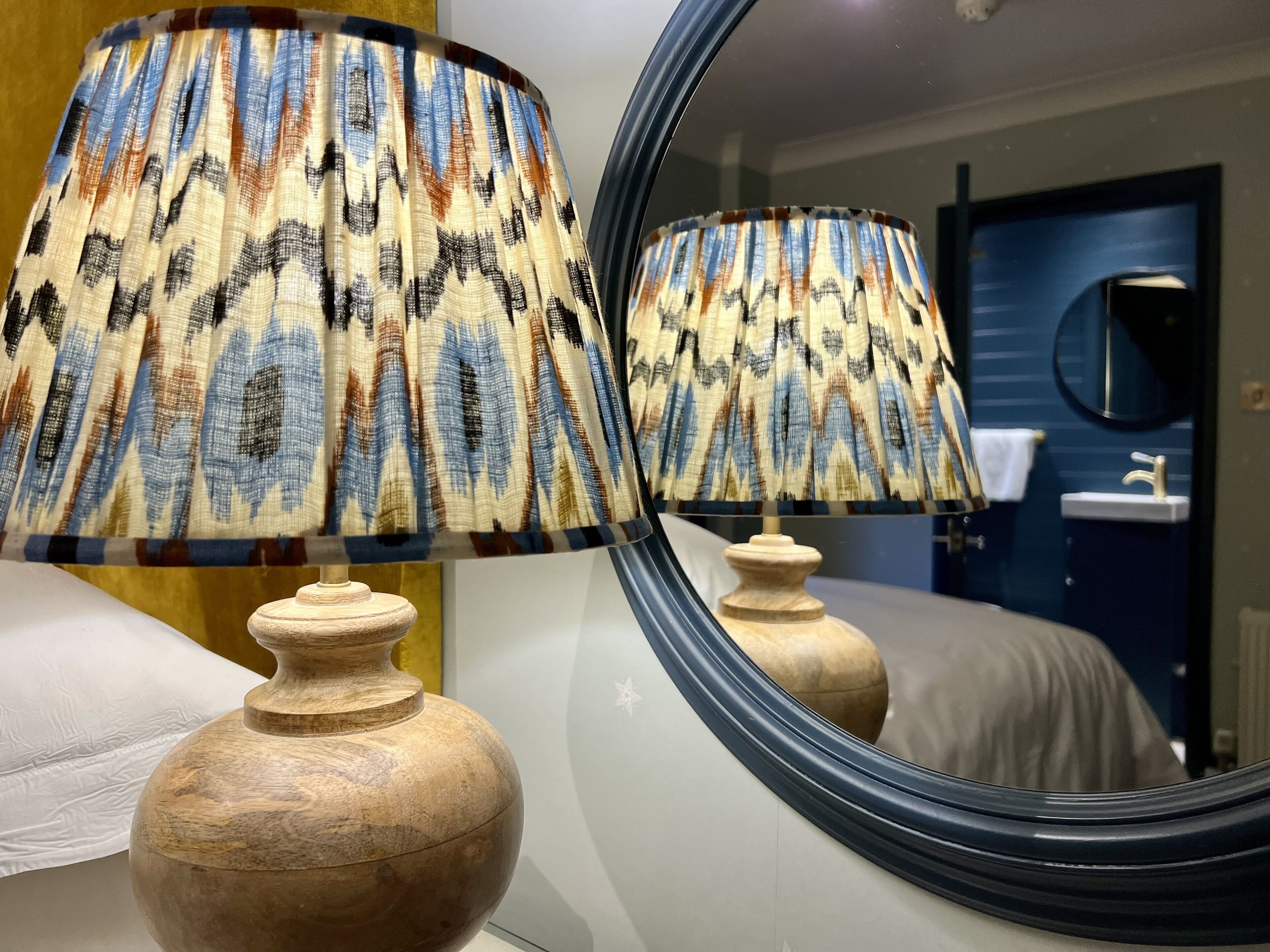 A room with a bed, a wooden nightstand, a mirror, and a bathroom visible through the mirror.