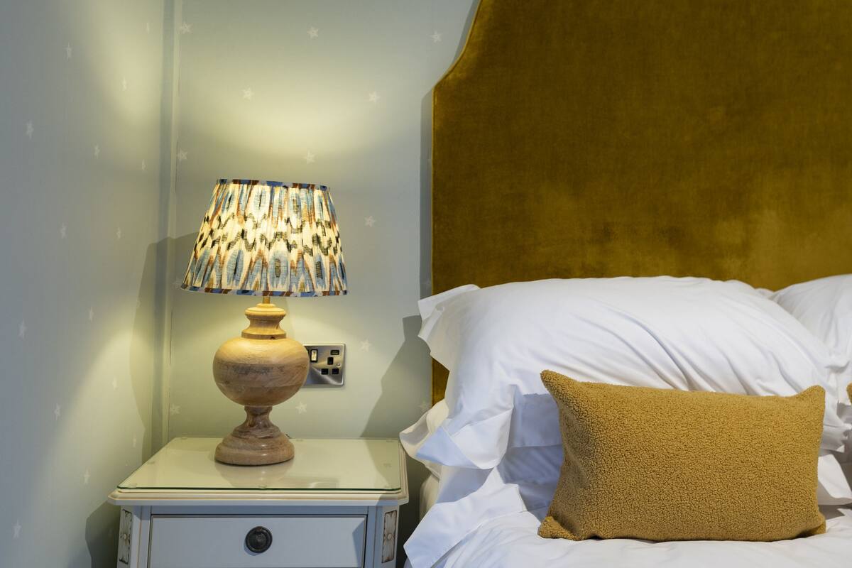 A neatly made bed with white linens and a single mustard-colored pillow. A wooden bedside table with a patterned lamp is positioned next to the bed.