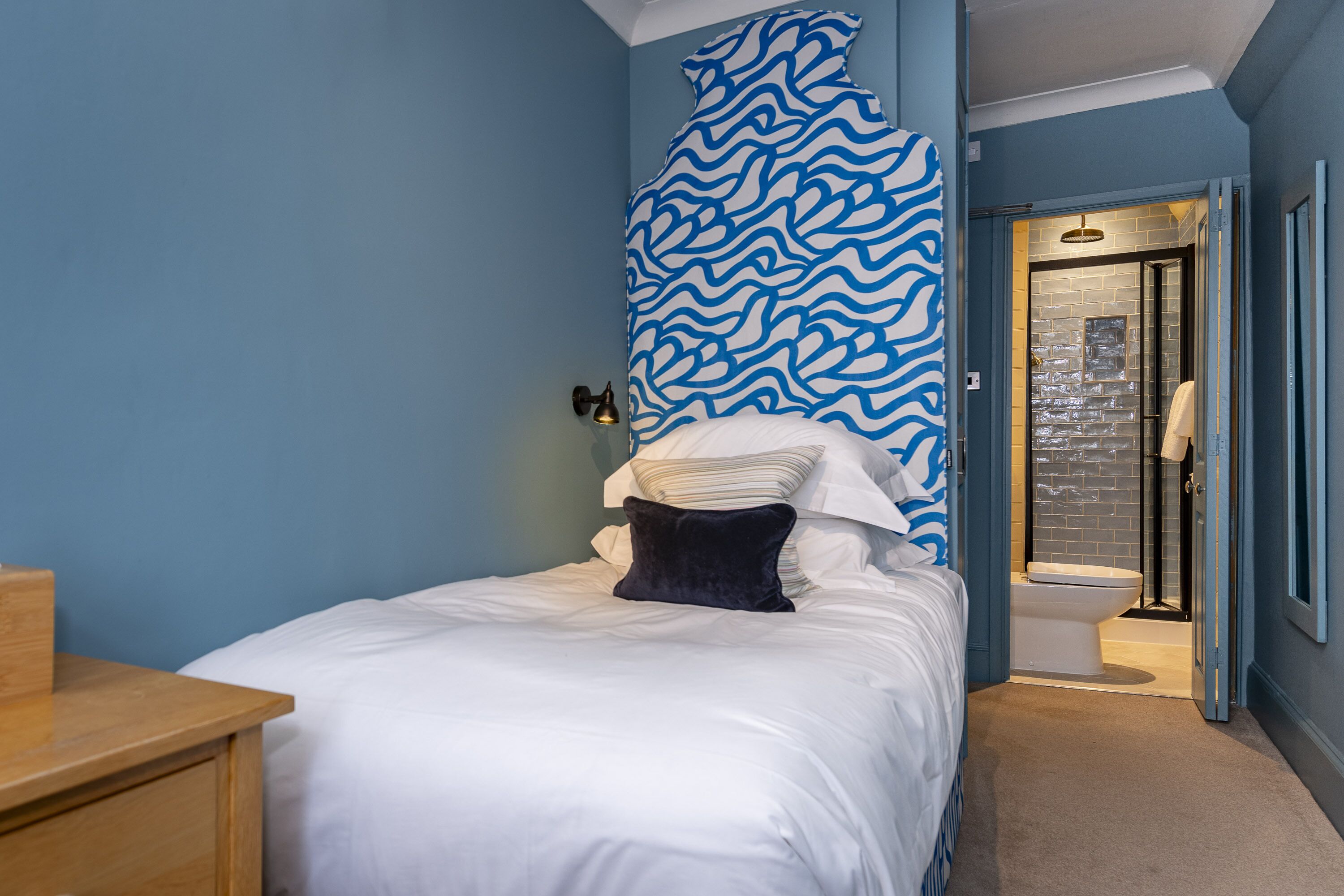 Un dormitorio con cama, una mesita de noche de madera, un muro con diseño ondulado en azul y un baño con cabina de ducha de vidrio.