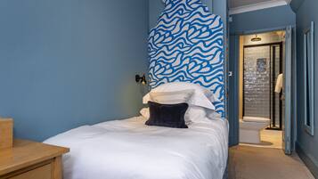 Un dormitorio con cama, una mesita de noche de madera, un muro con diseño ondulado en azul y un baño con cabina de ducha de vidrio.
