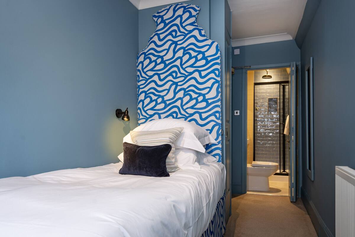 A bedroom with a bed, a blue and white patterned wall, a white radiator, and a bathroom visible through an open door.