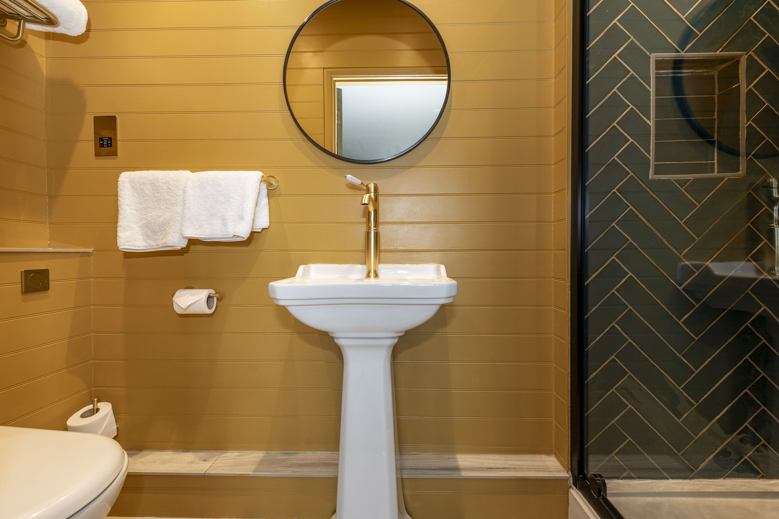 Un baño con lavamanos pedestal, espejo redondo y perchero.