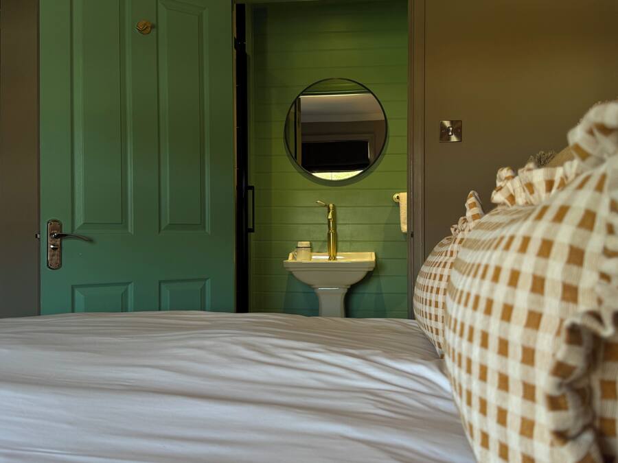 Une pièce avec une porte verte, un miroir rond au-dessus d’un lavabo et un lit recouvert d’une couverture à carreaux.