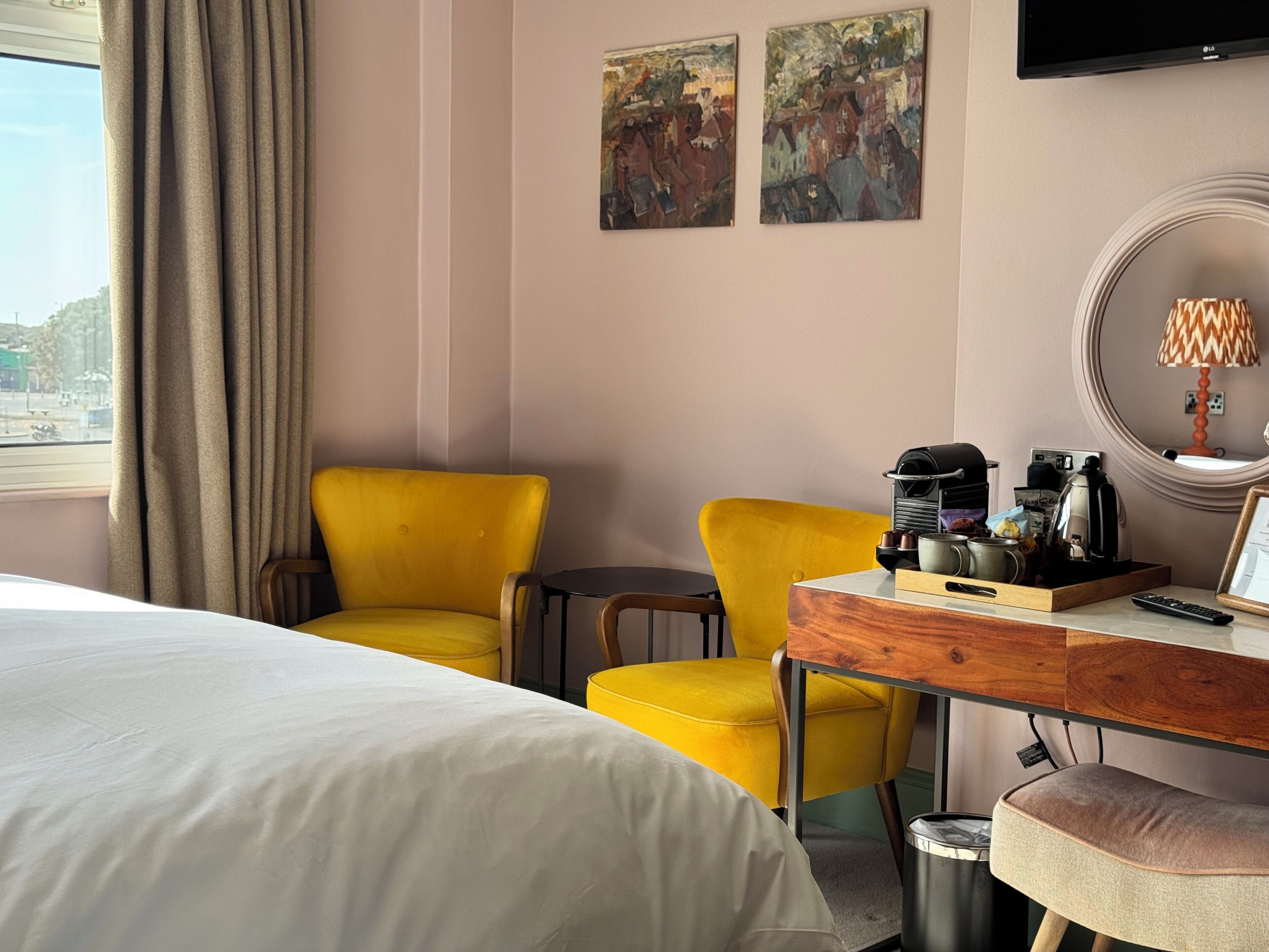 A hotel room with a bed, two yellow chairs, a wooden side table with a coffee maker, a TV, and artwork on the wall.