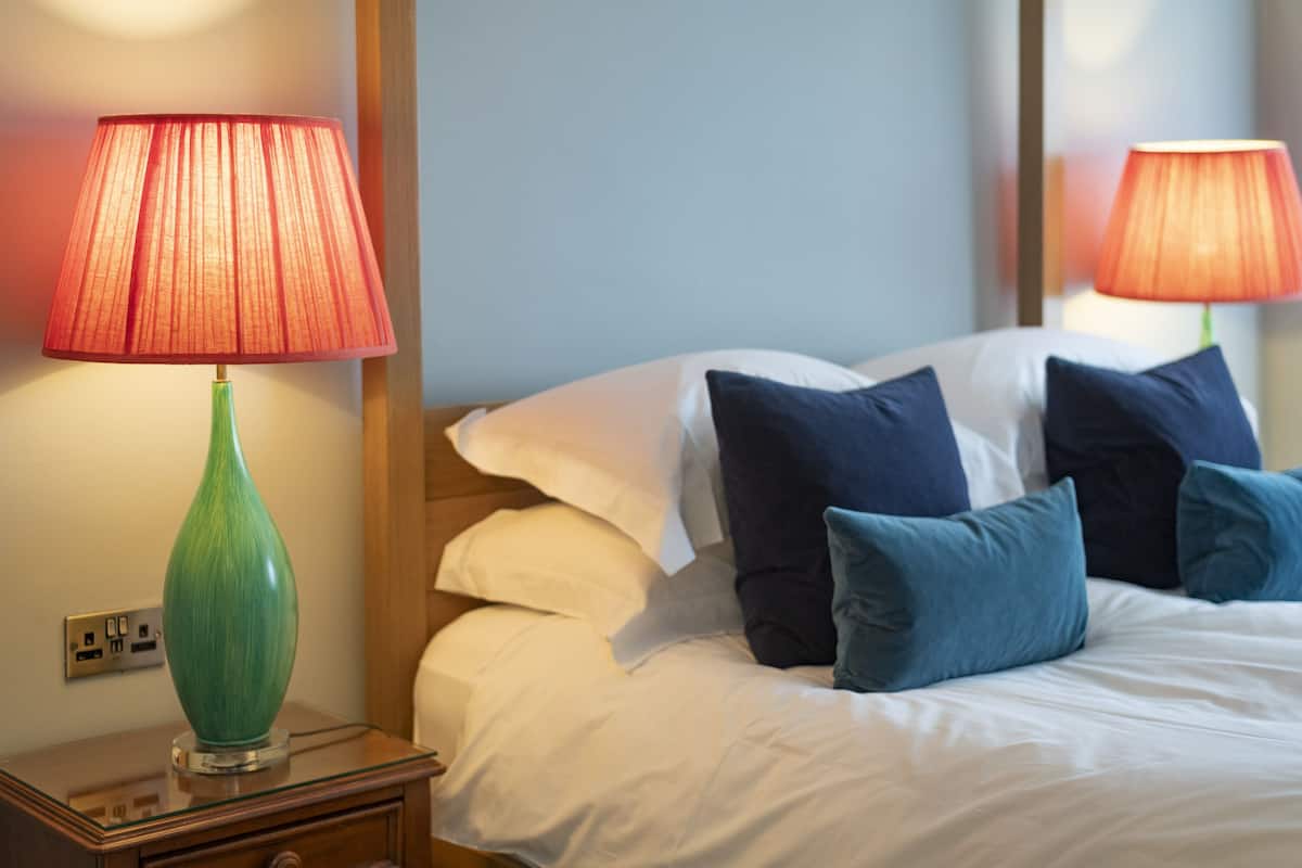 A neatly made bed with white and blue pillows, a green vase on a bedside table, and two lamps with red shades.