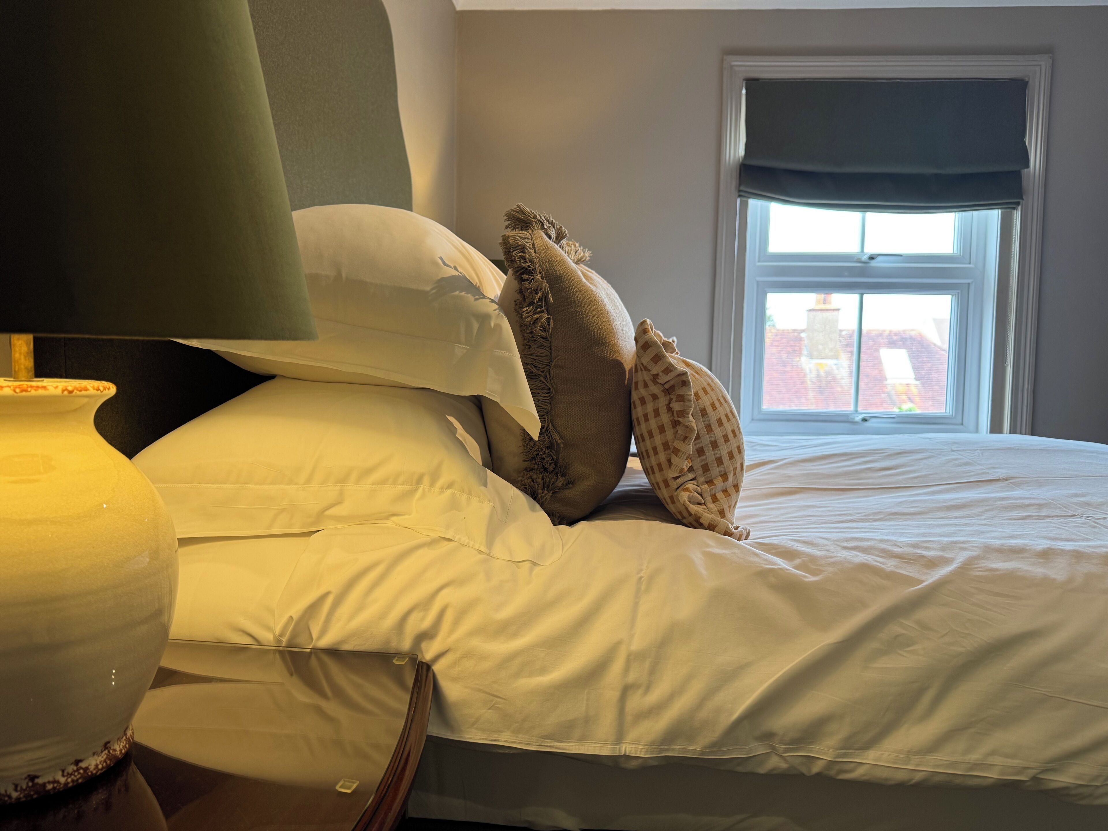 Una cama con ropa de cama blanca, cabecera verde y ventana con vistas a edificios.
