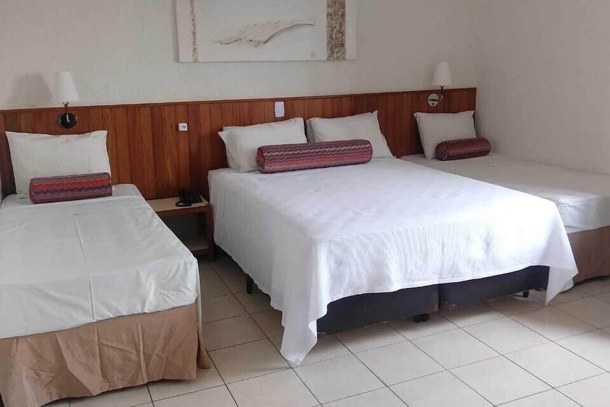 Quarto de hotel com três camas, cada uma com roupa de cama branca e cabeceira de madeira. Há uma mesa de cabeceira com abajur entre as camas, e um ar condicionado fixado na parede.