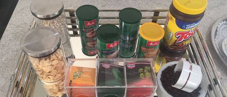 Una encimera de cocina con diversos alimentos y recipientes, entre ellos frascos de especias, un recipiente con granos de café y una bandeja con distintos productos alimenticios.