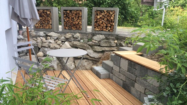 Una terraza de madera con un muro de piedra, una mesa pequeña y un espacio para sentarse.