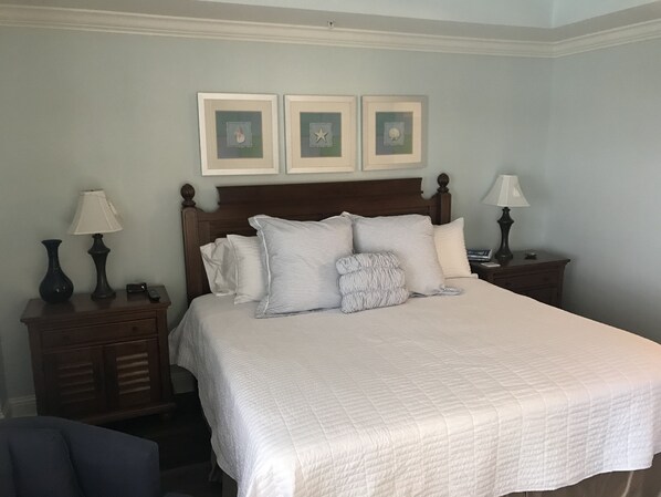 Un lit bien fait, avec une literie blanche et des oreillers à rayures, flanqué de deux tables de chevet surmontées de lampes, le tout adossé à un mur bleu clair orné de trois tableaux encadrés.