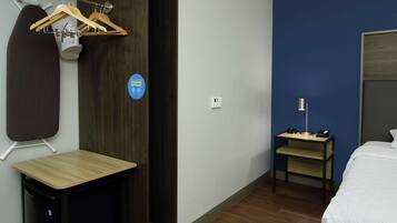 A hotel room with a bed, a desk, a chair, a coat rack, and a small table with a lamp.