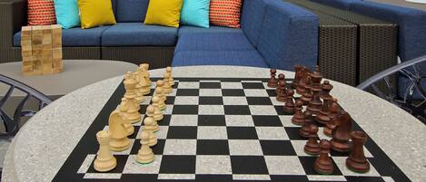 A checkered chessboard with wooden pieces on a round table, set outdoors with a blue sofa and colorful cushions in the background.