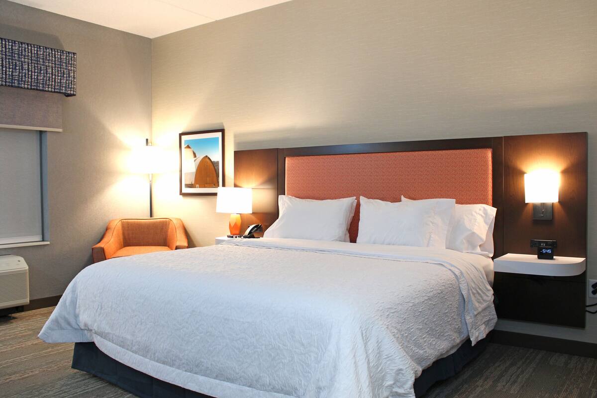 A hotel room with a large bed, two bedside tables, a chair, a wall-mounted air conditioner, and a framed picture on the wall.