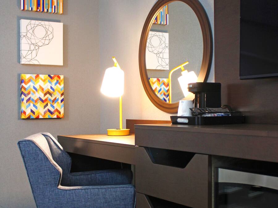 A hotel room with a desk, chair, and a large mirror. There are two artworks on the wall and a coffee machine on the desk.
