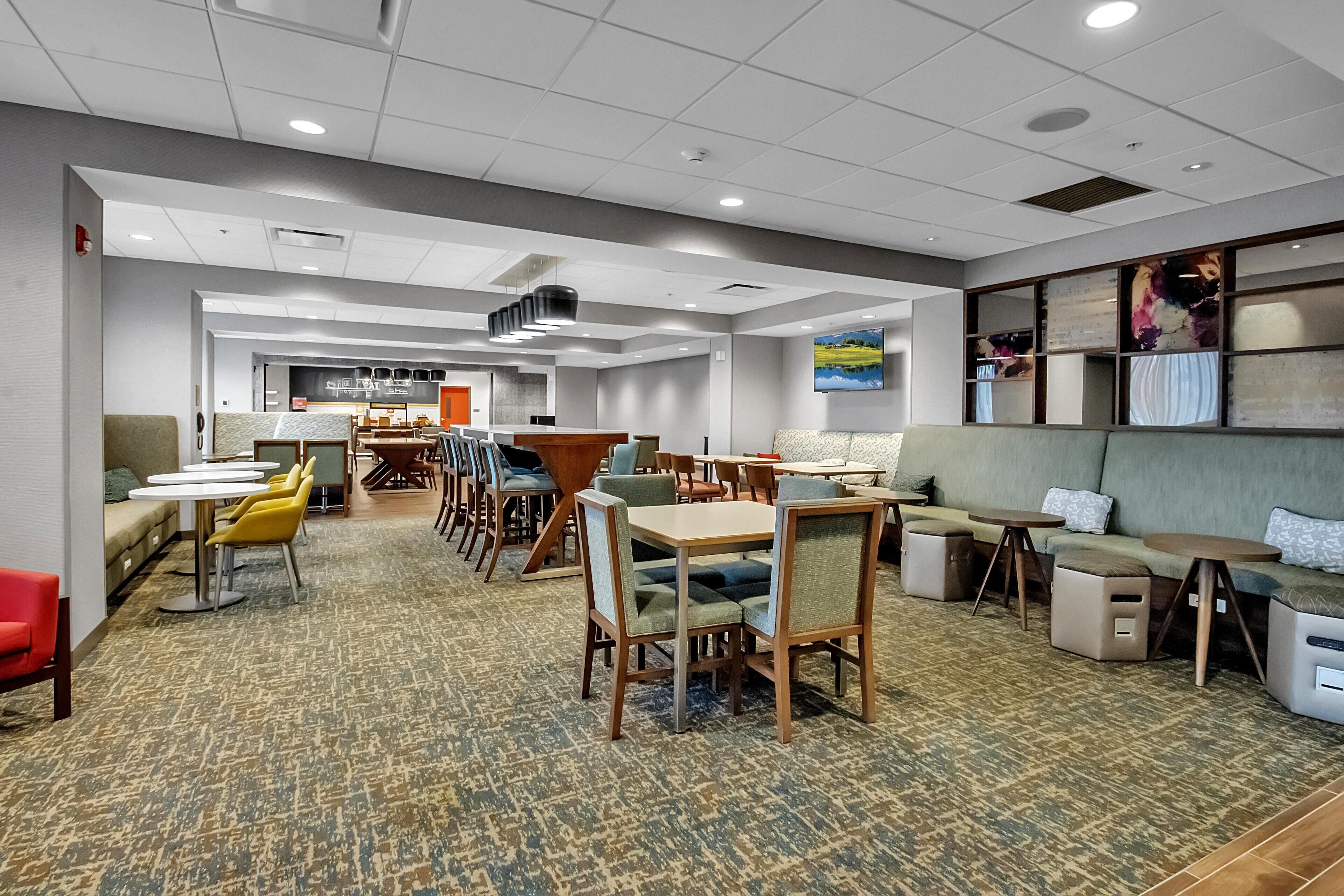 A hotel lobby with multiple seating areas, including tables and sofas, carpeted flooring, and a ceiling with recessed lighting.