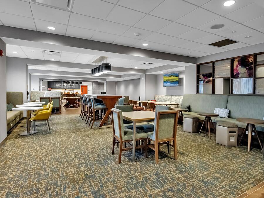 A hotel lobby with multiple seating areas, including tables and sofas, carpeted flooring, and a ceiling with recessed lighting.