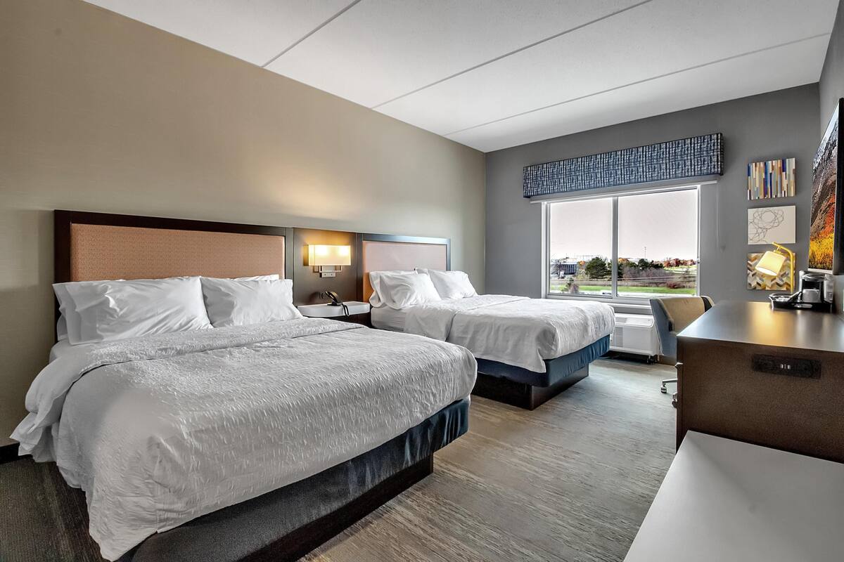 A hotel room with two beds, a desk, a window with a city view, and artwork on the walls.